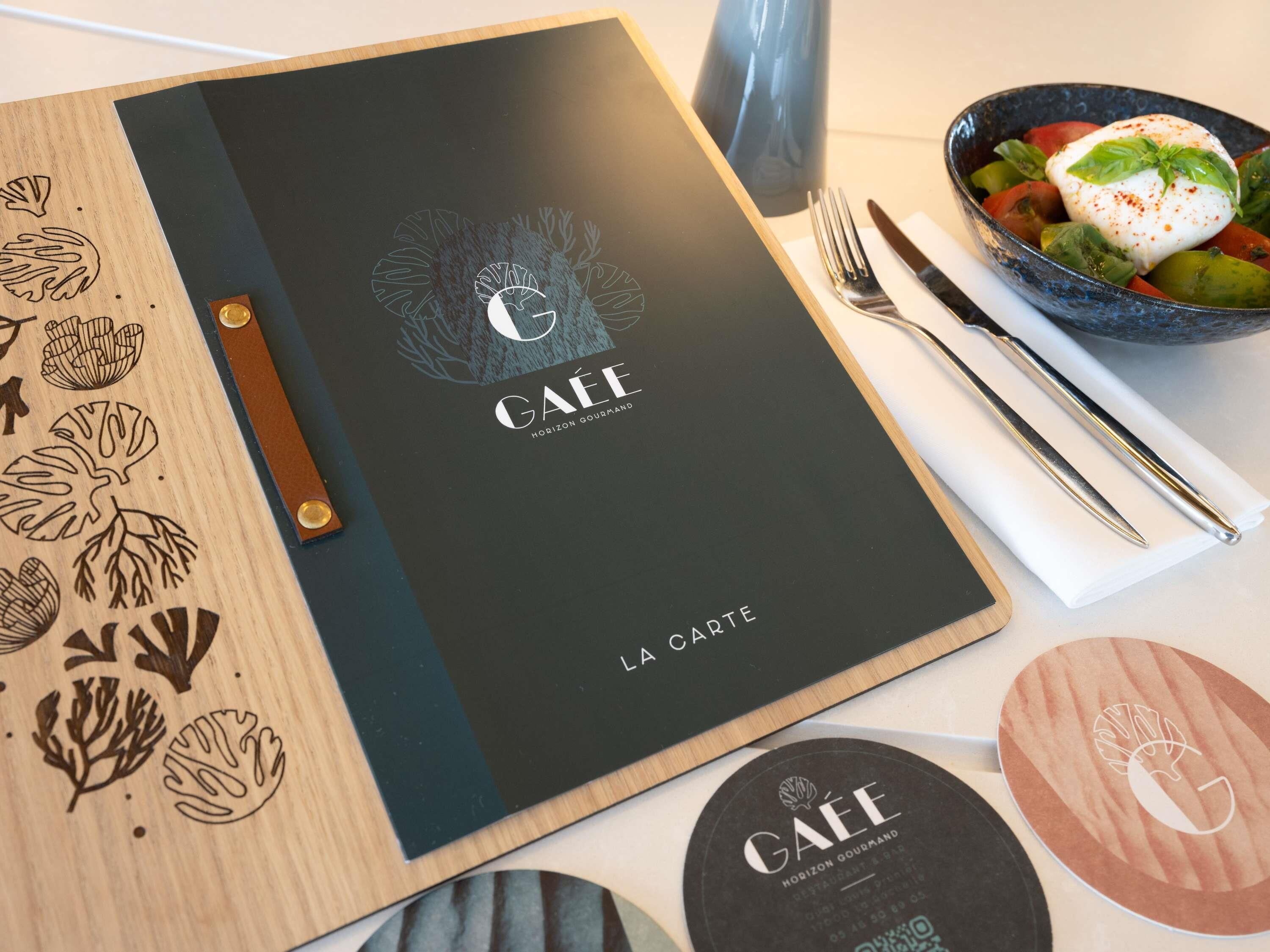 Un panel de menú con la marca 'GAËE', un aparador de madera con diseños de hojas y un tazón de comida con cobertura blanca.
