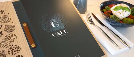 Un panel de menú con la marca 'GAËE', un aparador de madera con diseños de hojas y un tazón de comida con cobertura blanca.