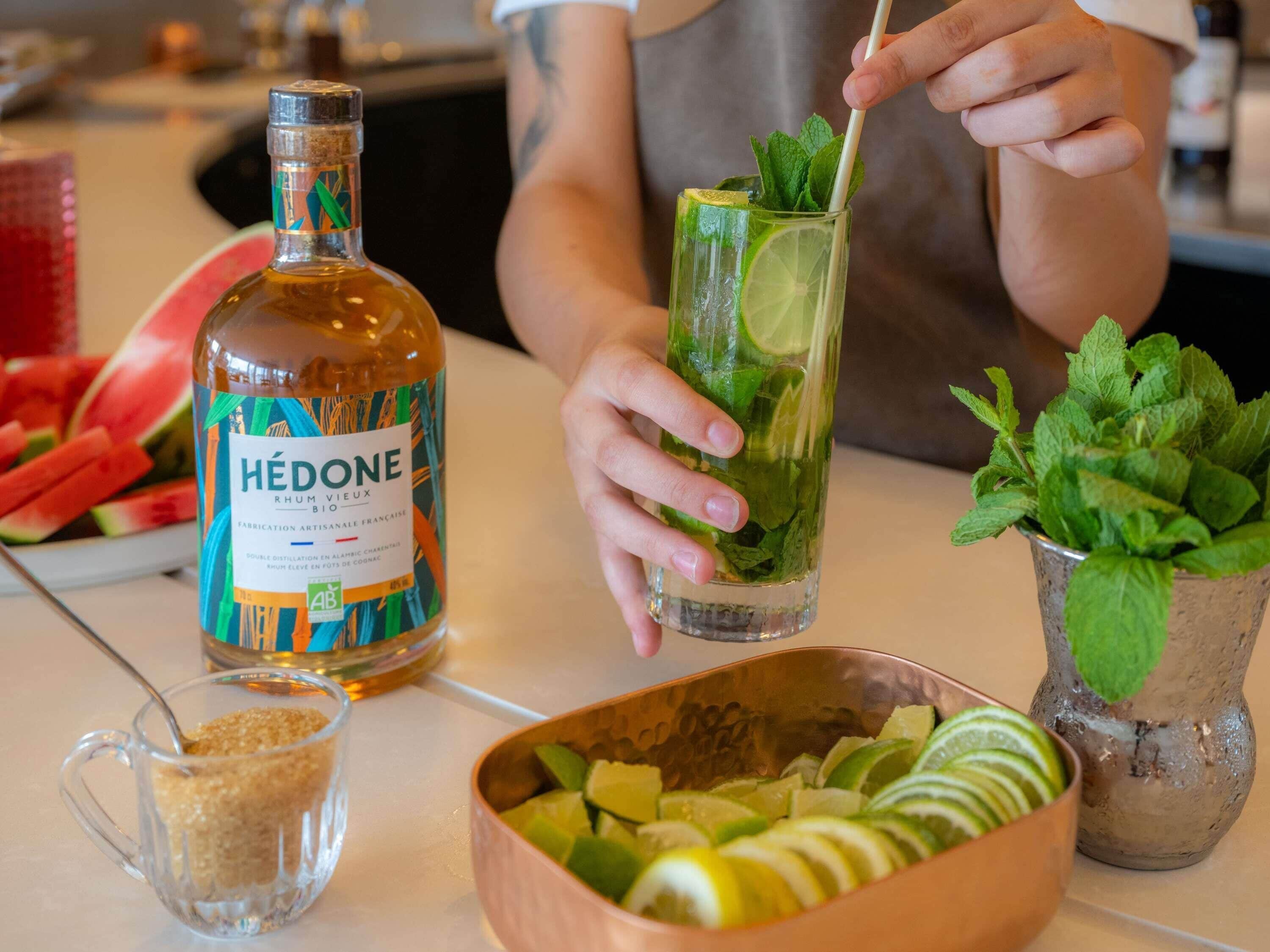 Una persona preparando una bebida con una botella etiquetada 'HEDONE' y un vaso con lima y menta.