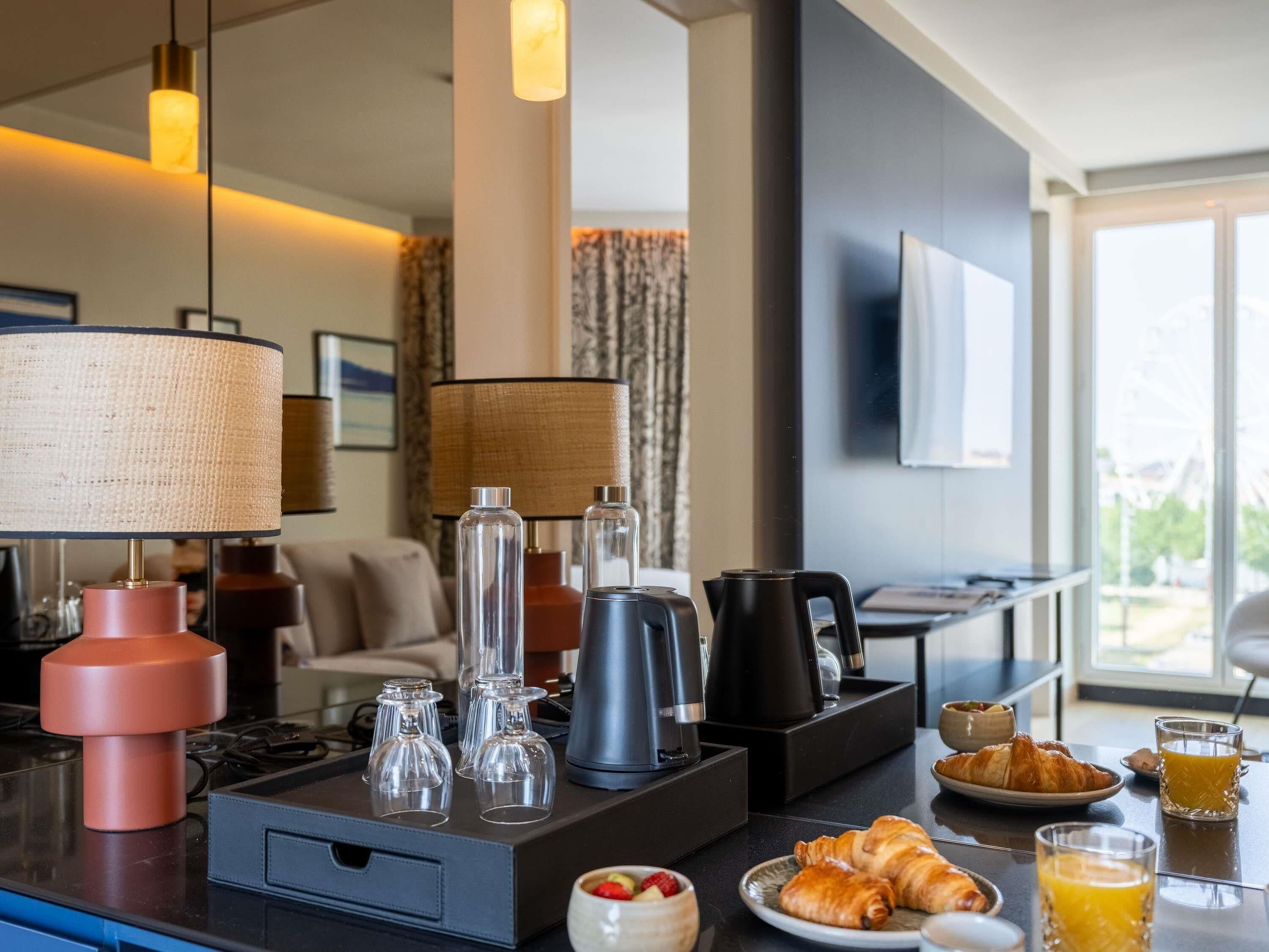 Una habitación de hotel moderna con zona de comedor, cafetera, tetera y productos para el desayuno, como croissants y jugo.