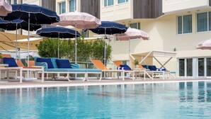 Un espace piscine avec des chaises longues et des parasols, une piscine à l’eau claire, et un bâtiment avec des fenêtres et des balcons en arrière-plan.