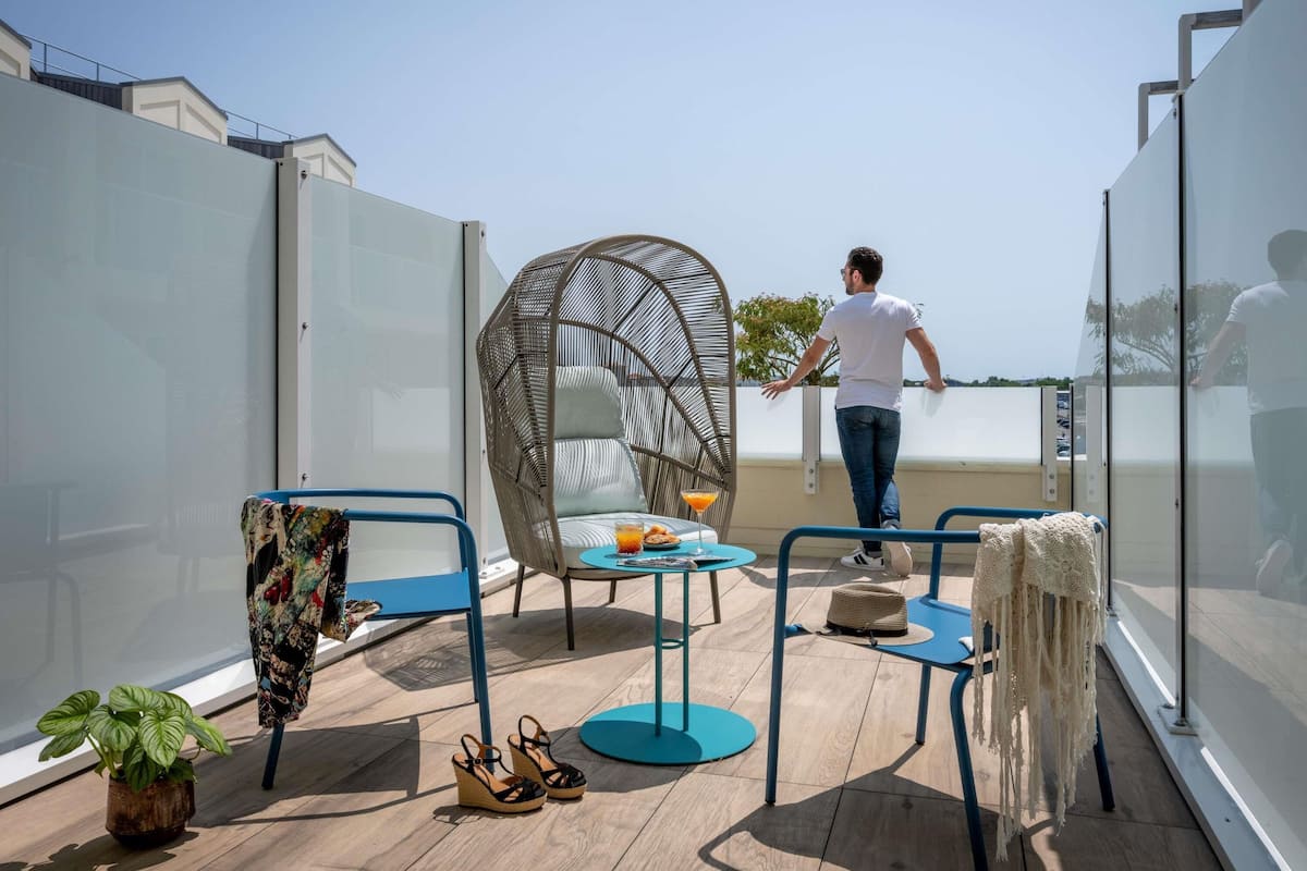 Una terraza en la azotea con una silla de mimbre, una mesita con una bebida, una planta en maceta y un par de zapatos.