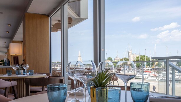 Une table à manger, agrémentée de verres à vin et d’une plante, donnant sur une marina où sont amarrés des bateaux et où se dresse une tour.