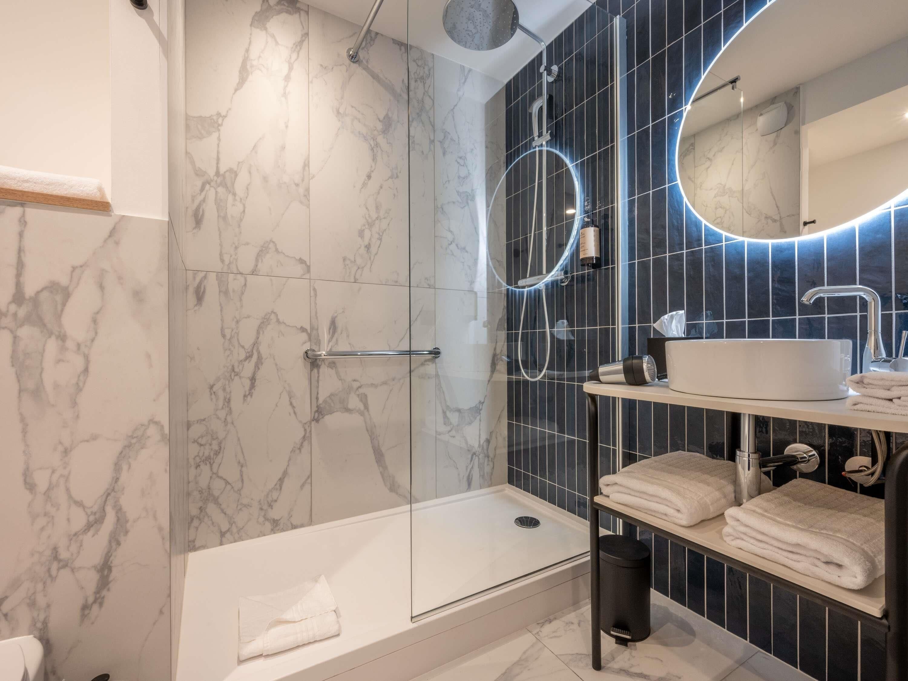 A modern bathroom with a glass-enclosed shower, a white sink, and a mirror with a circular light.