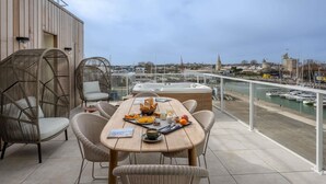 Une terrasse meublée de mobilier en osier, une table en bois où est servi le petit-déjeuner, et une vue sur un port avec des bateaux et des bâtiments.