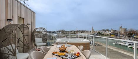 Una terraza con mobiliario de mimbre, una mesa de madera con desayuno, y vistas a un puerto con barcos y edificios.