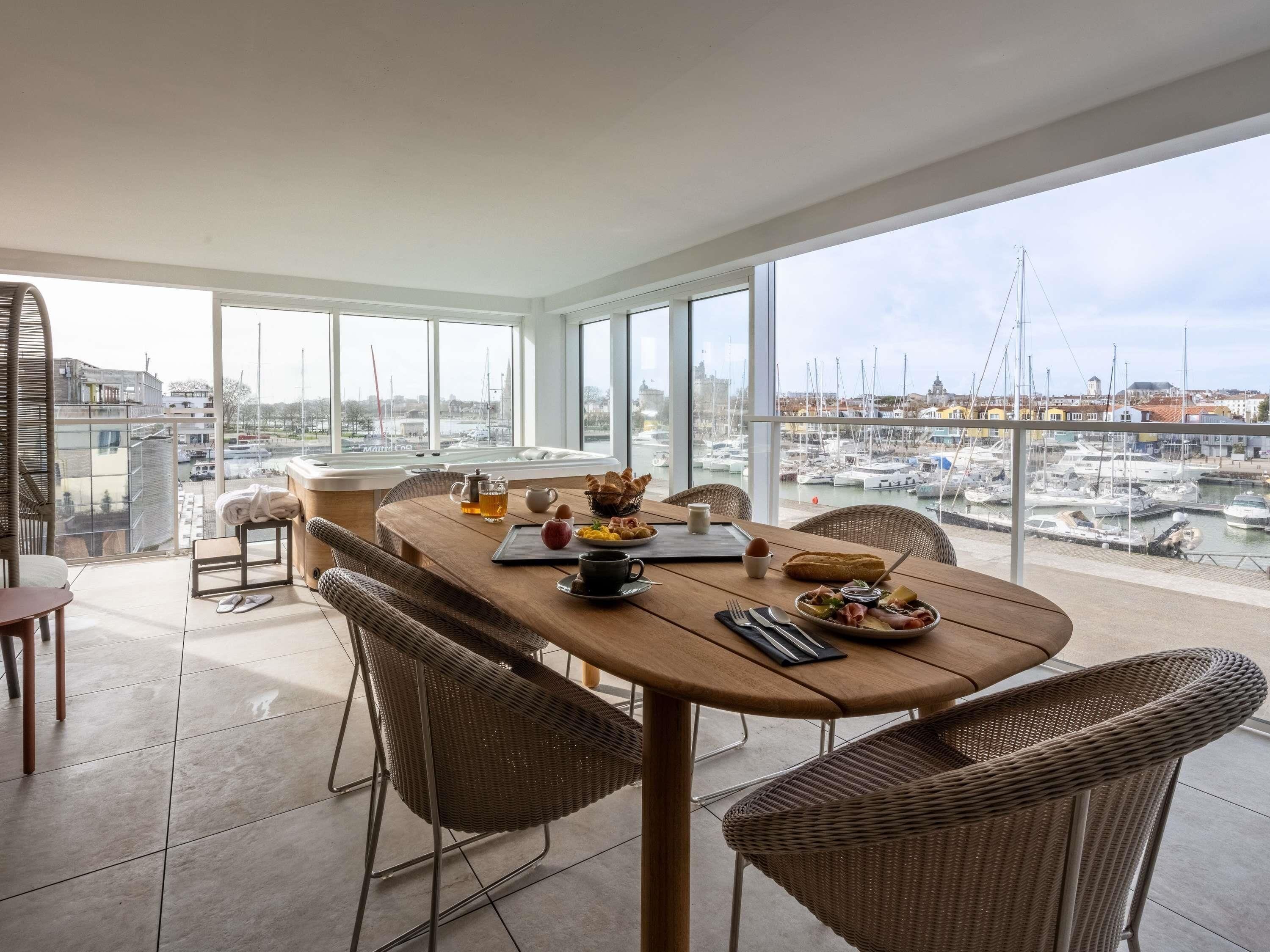 A dining area with a view of a marina, wicker chairs, a wooden table, and breakfast items.