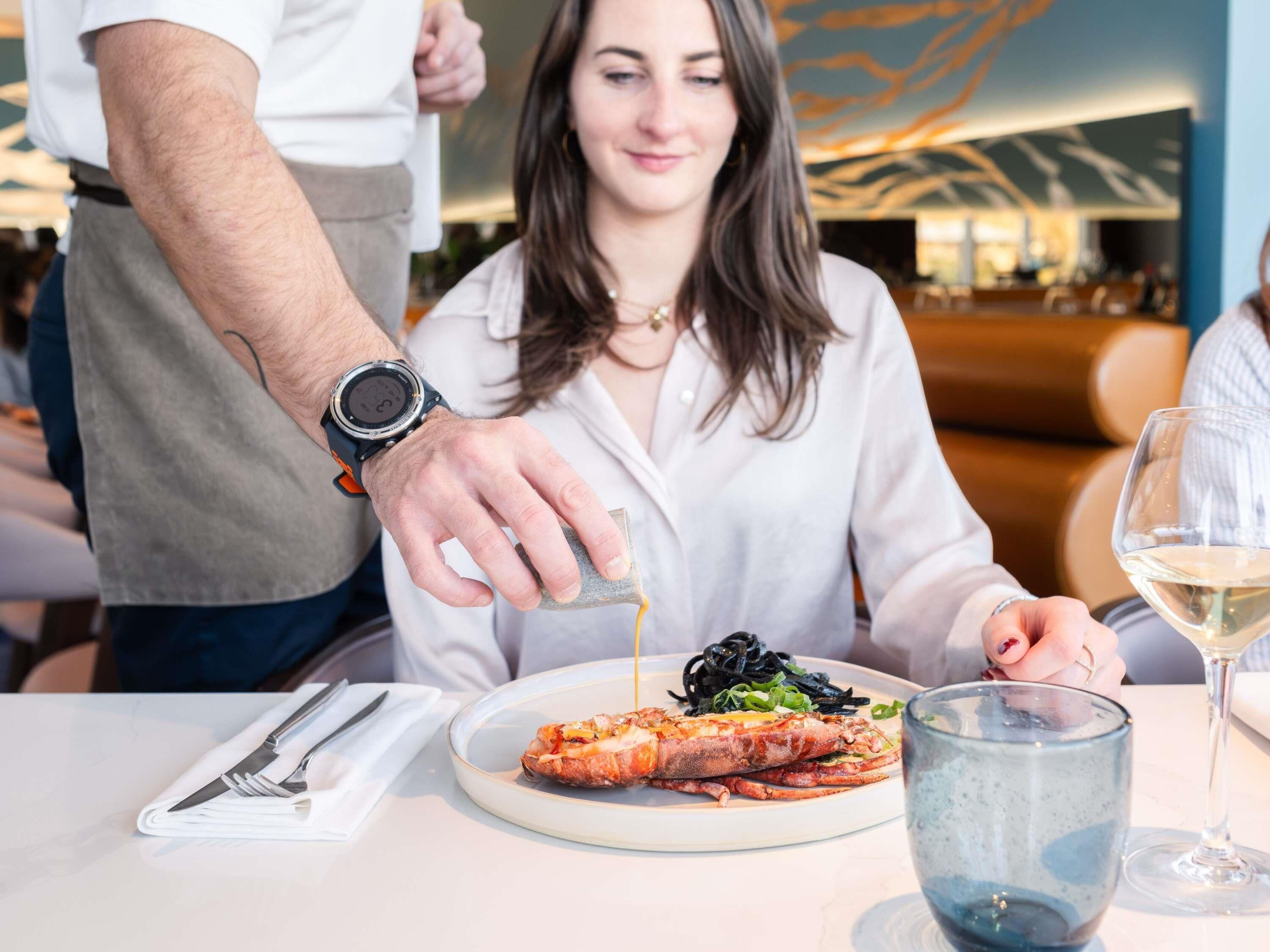 A dining table with a plate of food, a glass of wine, and a watch on a wrist.