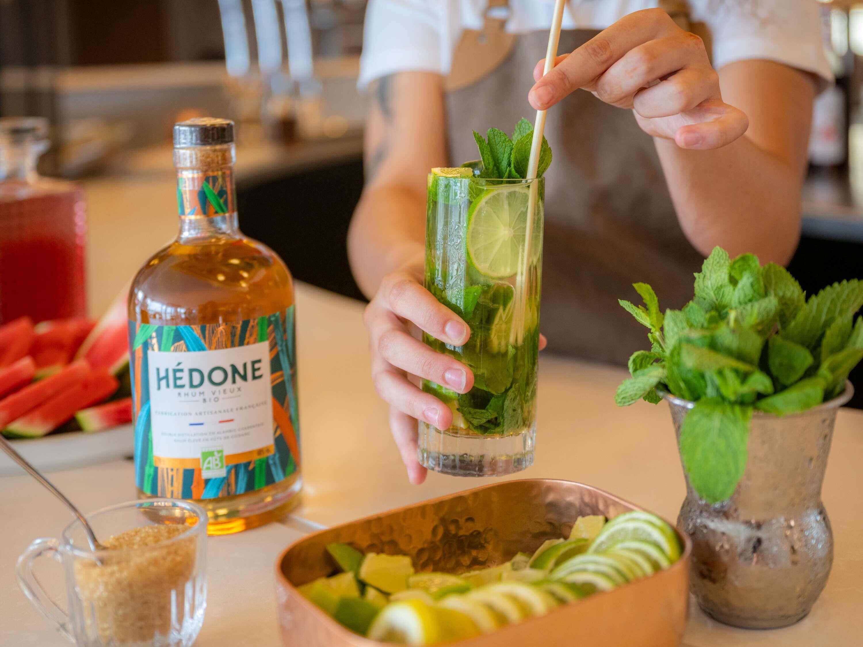 A person preparing a drink with a bottle labeled 'Hédone' and a bowl of sliced fruits.