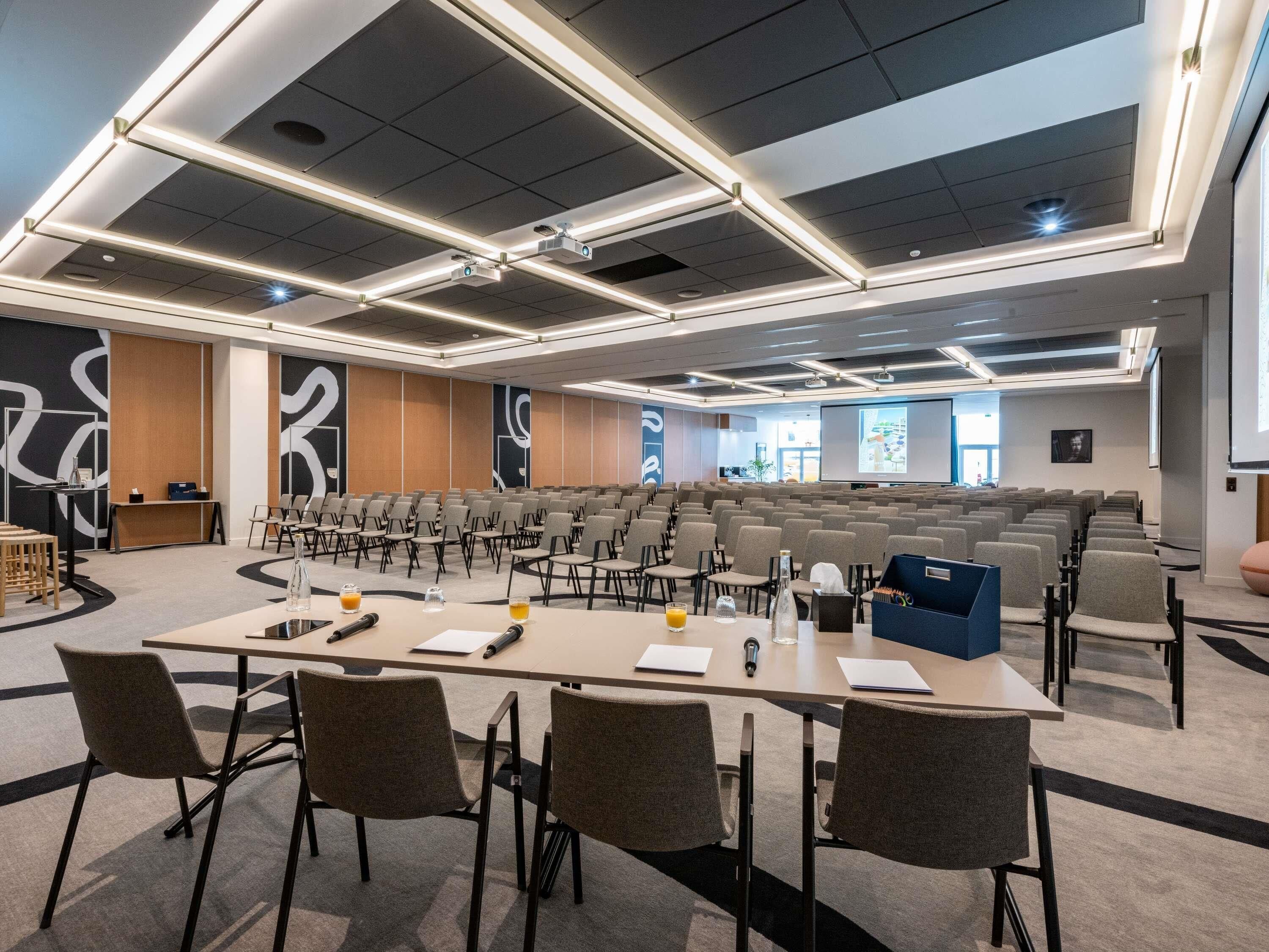 A conference room with rows of chairs, a long table with chairs, a projector, and a screen.