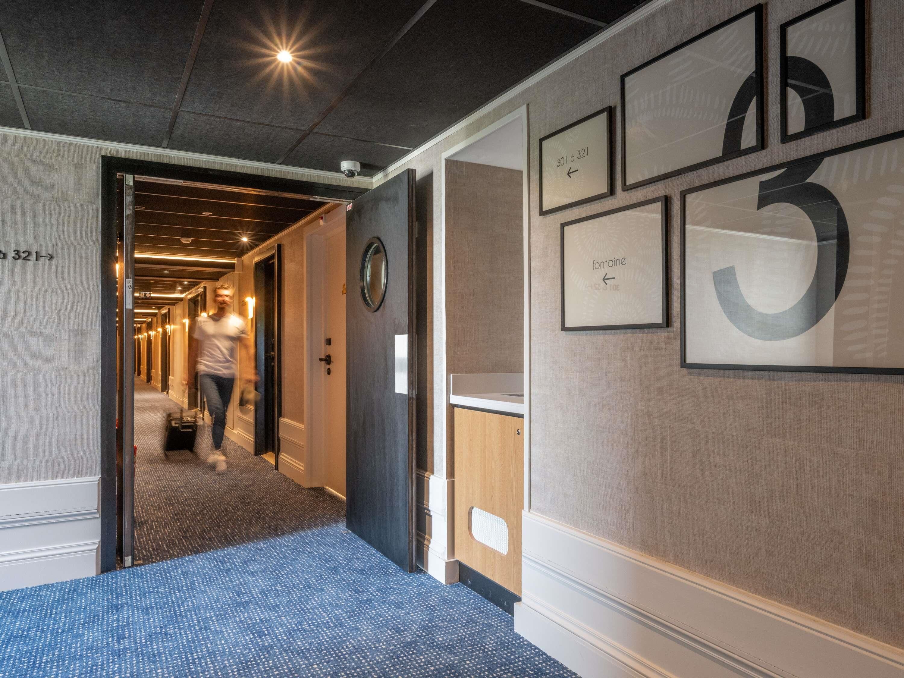 Un couloir d’hôtel avec des œuvres d’art encadrées, une moquette bleue et une personne qui marche.