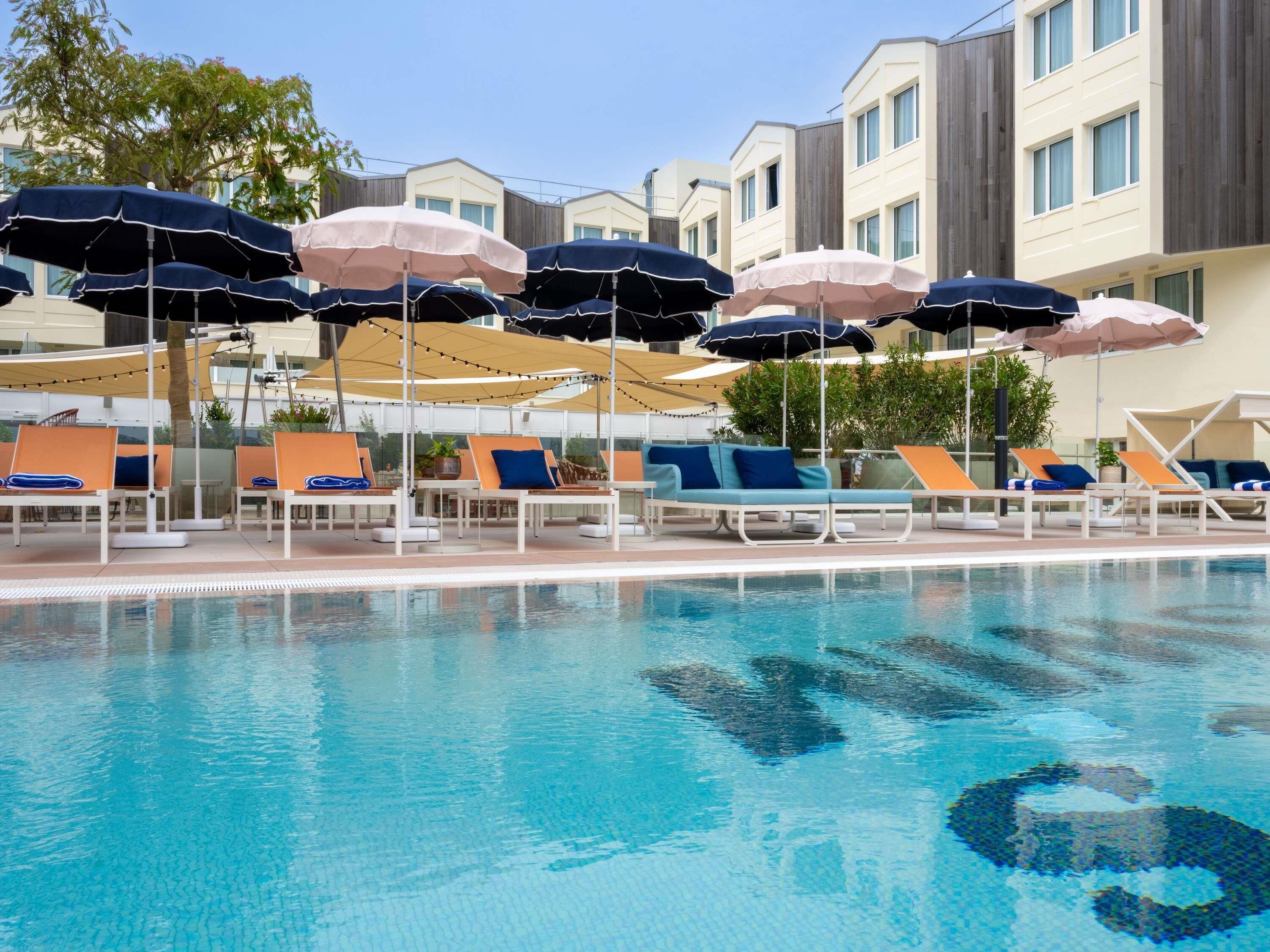Zona junto a la piscina con sillones de descanso y sombrillas, una piscina cristalina y edificios modernos al fondo.