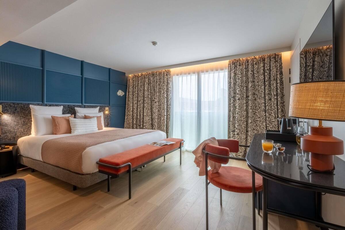 Une chambre d’hôtel moderne avec un grand lit, un bureau avec une lampe, une chaise et une petite table avec une boisson.
