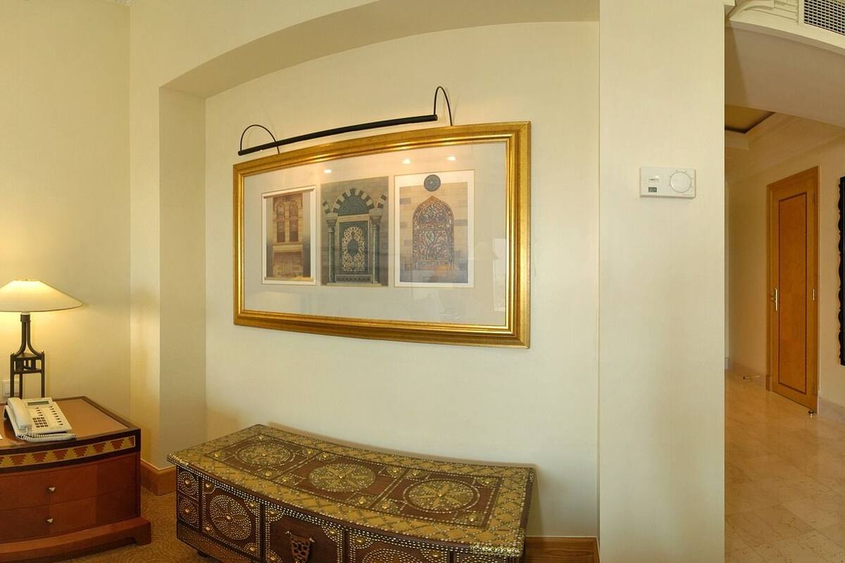 Quarto de hotel com uma cama grande, guarda-roupa de madeira, escrivaninha com cadeira, mesinha com abajur e um aparador decorativo.