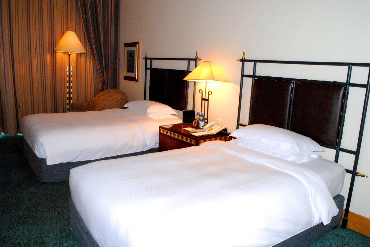 Quarto de hotel com duas camas de solteiro, mesa de cabeceira com abajur, telefone e pequena mesa com garrafa e copos.