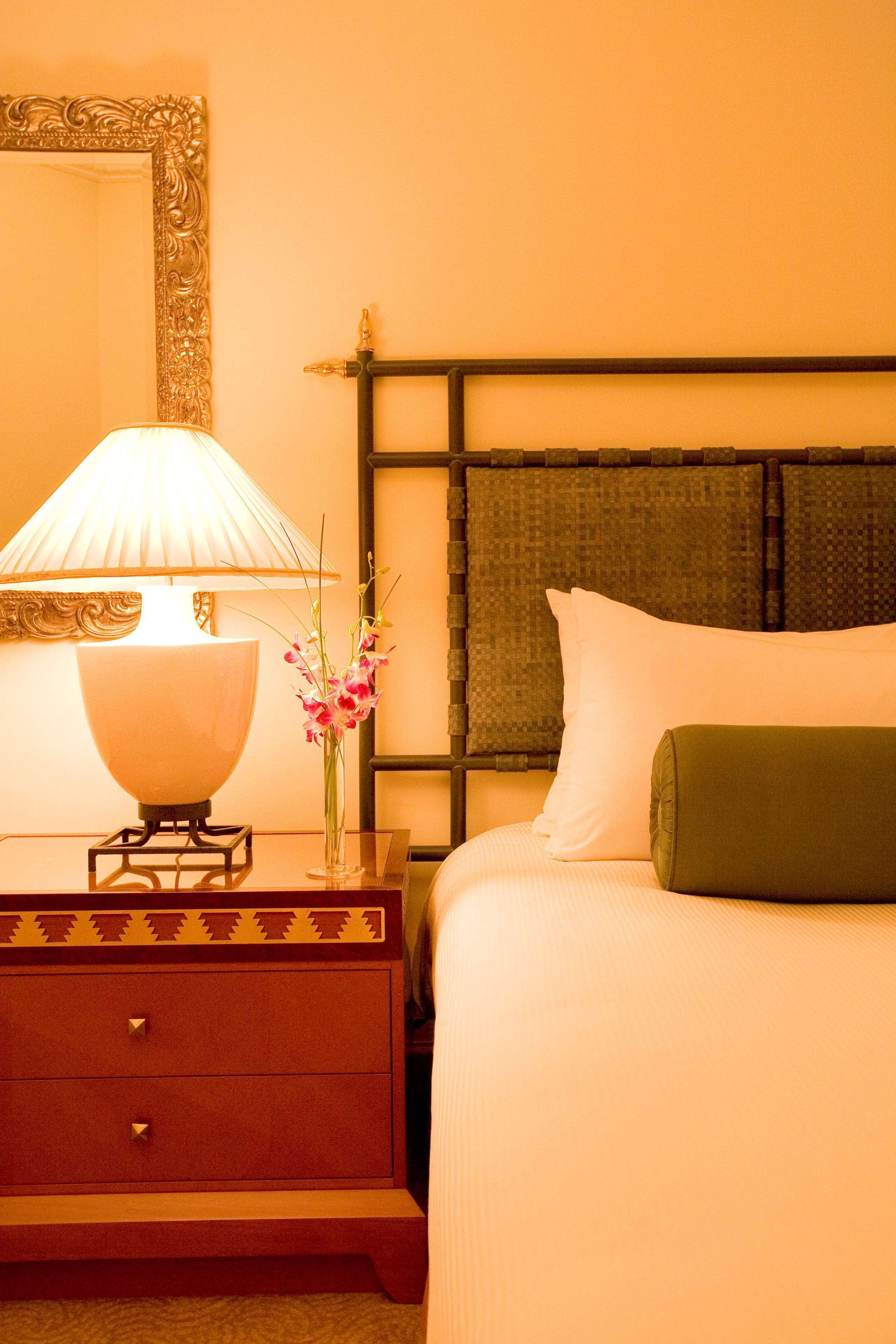 A hotel room with a bed, a nightstand with a lamp, a vase with flowers, and a mirror with an ornate frame.