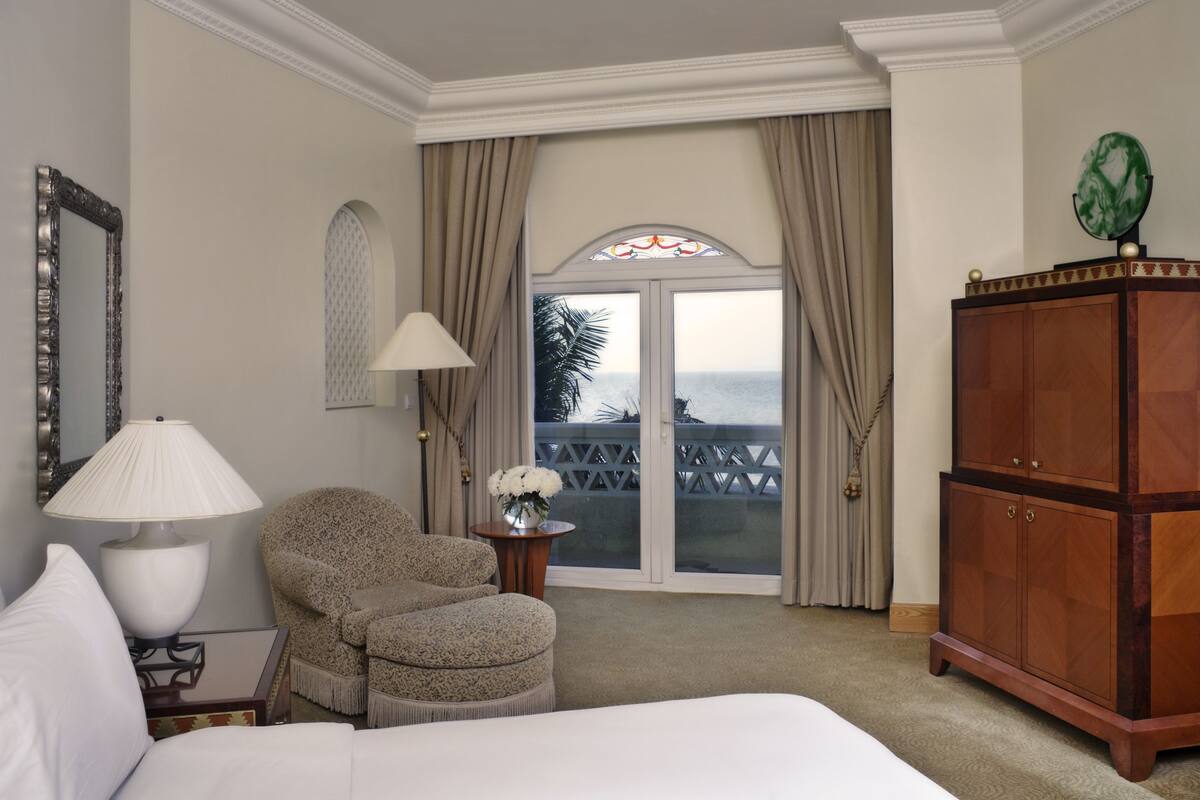 Quarto de hotel com cama, cadeira, luminária, armário e vista do oceano por uma porta de vidro.