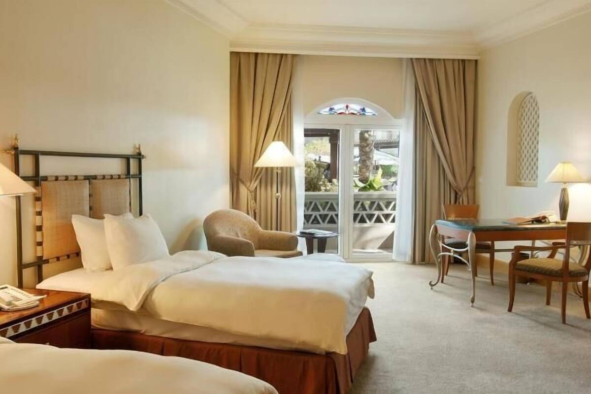 Quarto de hotel com duas camas, uma escrivaninha, uma cadeira e uma varanda com cortinas.