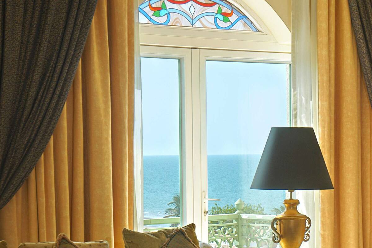 Uma sala de estar aconchegante, com um sofá, uma mesa de centro de vidro e uma luminária. Uma janela com cortinas revela uma vista para o oceano.