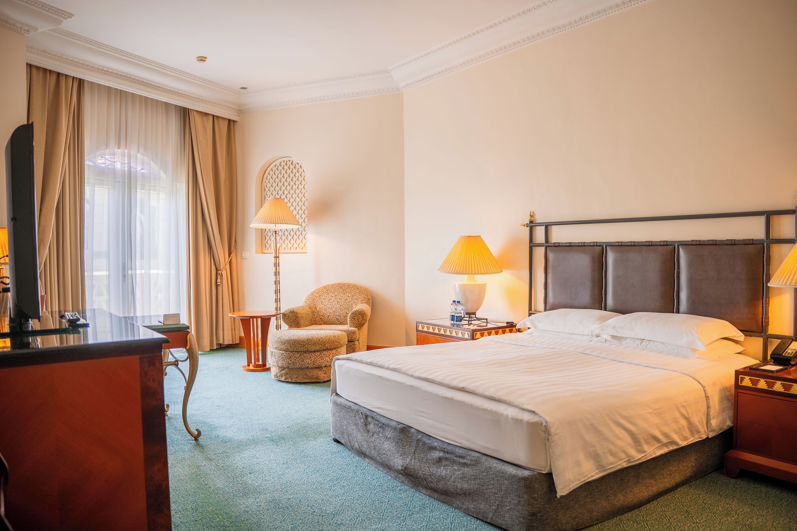 Habitación de hotel con cama, escritorio, silla, dos lámparas y un ventanal con cortinas.