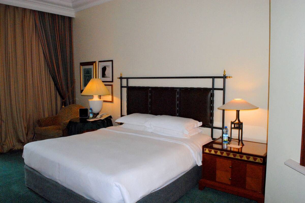 Quarto de hotel com cama grande, duas luminárias de cabeceira, mesa de cabeceira, uma cadeira e cortina.