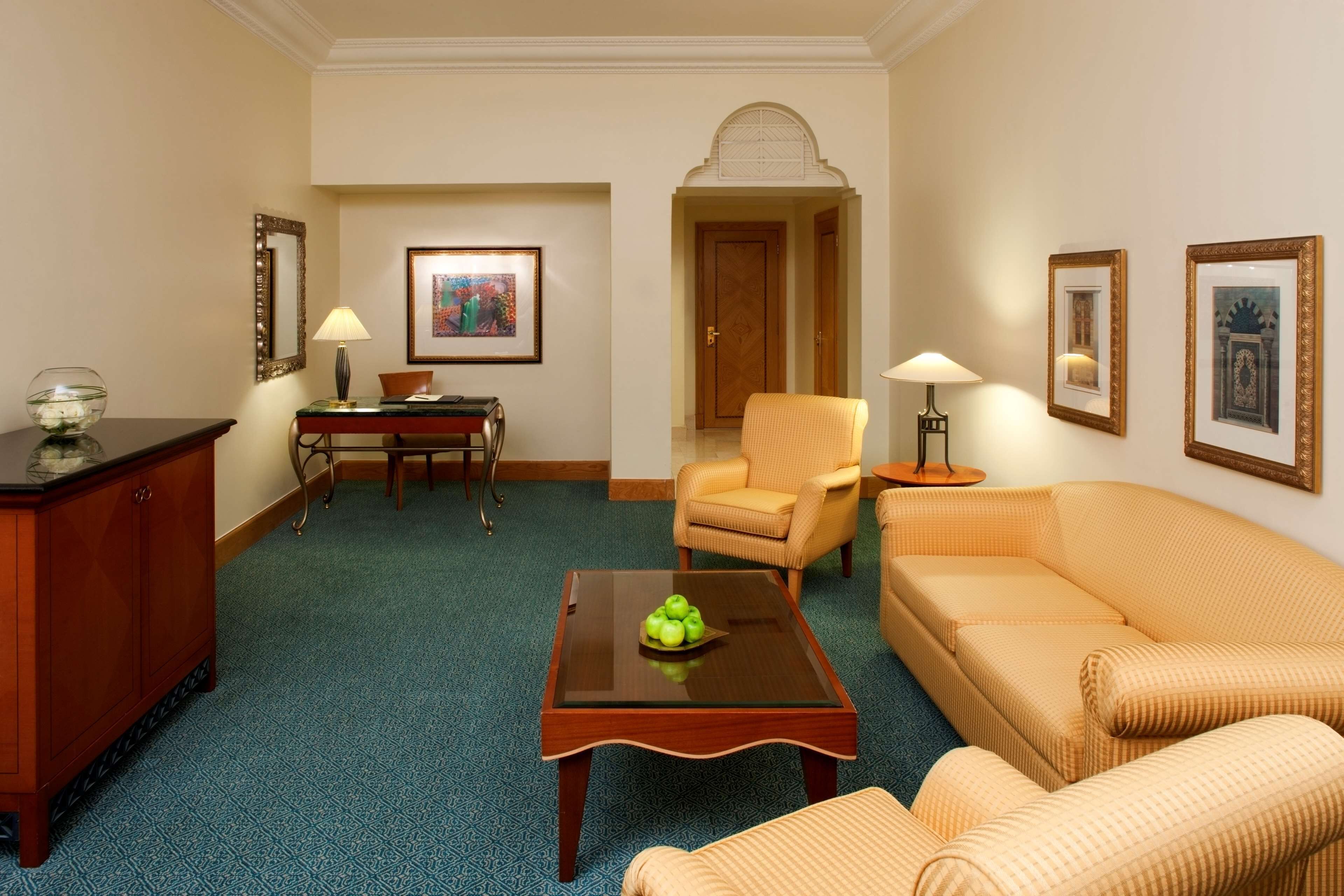 Habitación de hotel con alfombra azul, una mesa de centro de madera con manzanas, un sofá, una silla, un escritorio con lámpara y cuadros en las paredes.