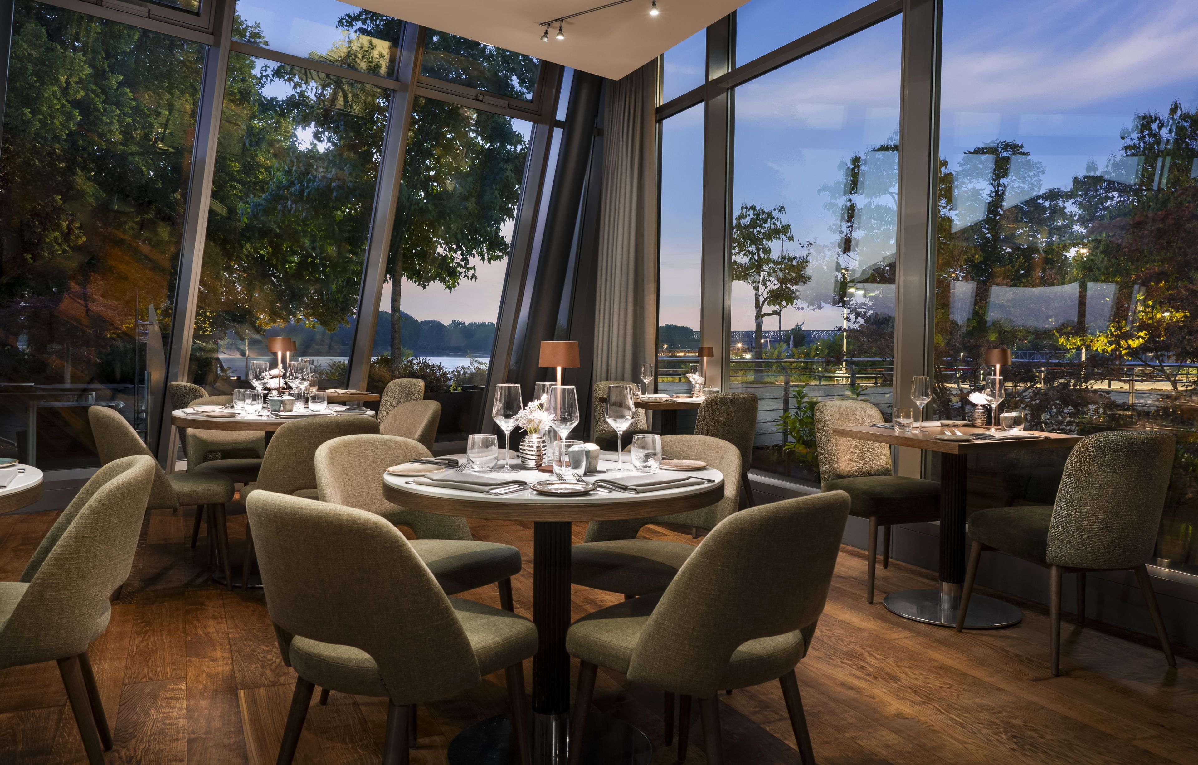 A restaurant with round tables and chairs, large windows, and a view of trees and a body of water.