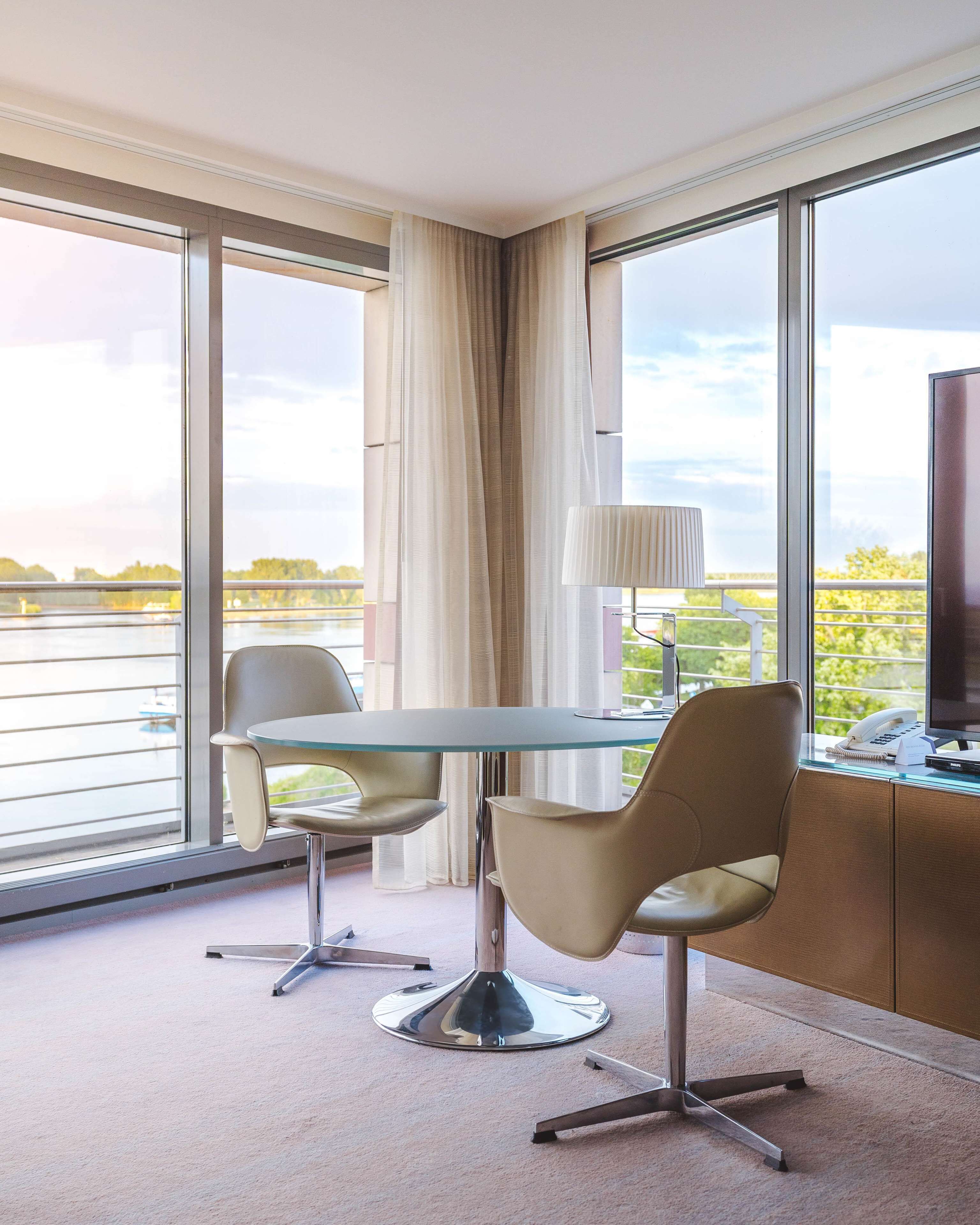 A modern hotel room with a glass table, two chairs, a lamp, a desk with a phone, and large windows offering a view of a body of water.