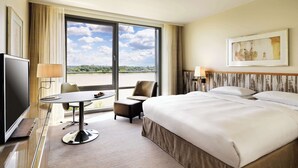 A hotel room with a large bed, a desk with a chair, a television, and a view of a body of water through a large window.