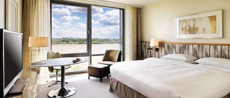 A hotel room with a large bed, a desk with a chair, a television, and a view of a body of water through a large window.