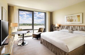 A hotel room with a large bed, a desk with a chair, a television, and a view of a body of water through a large window.