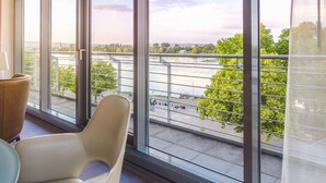A balcony with large windows offering a view of a river and greenery. There are two chairs and a table on the balcony.
