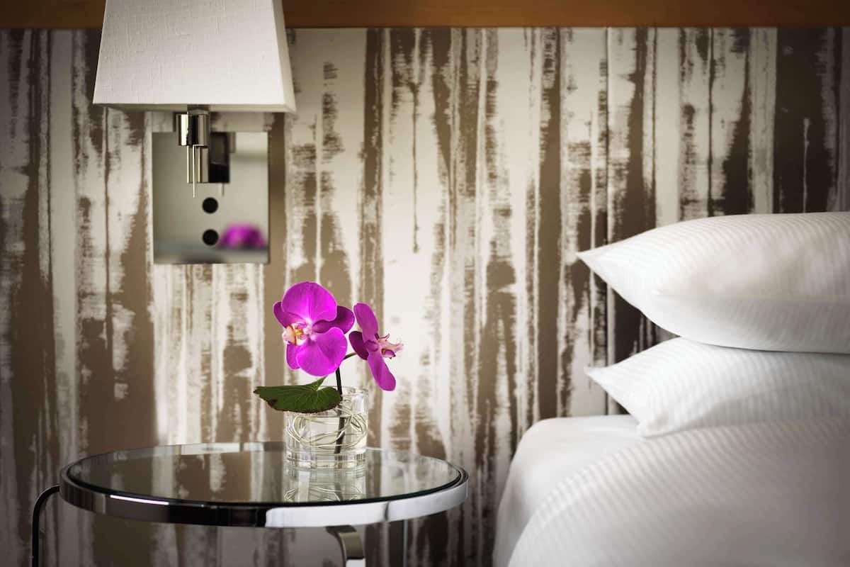 A hotel room with a bed, a glass table with a vase of purple flowers, and a wall-mounted lamp.