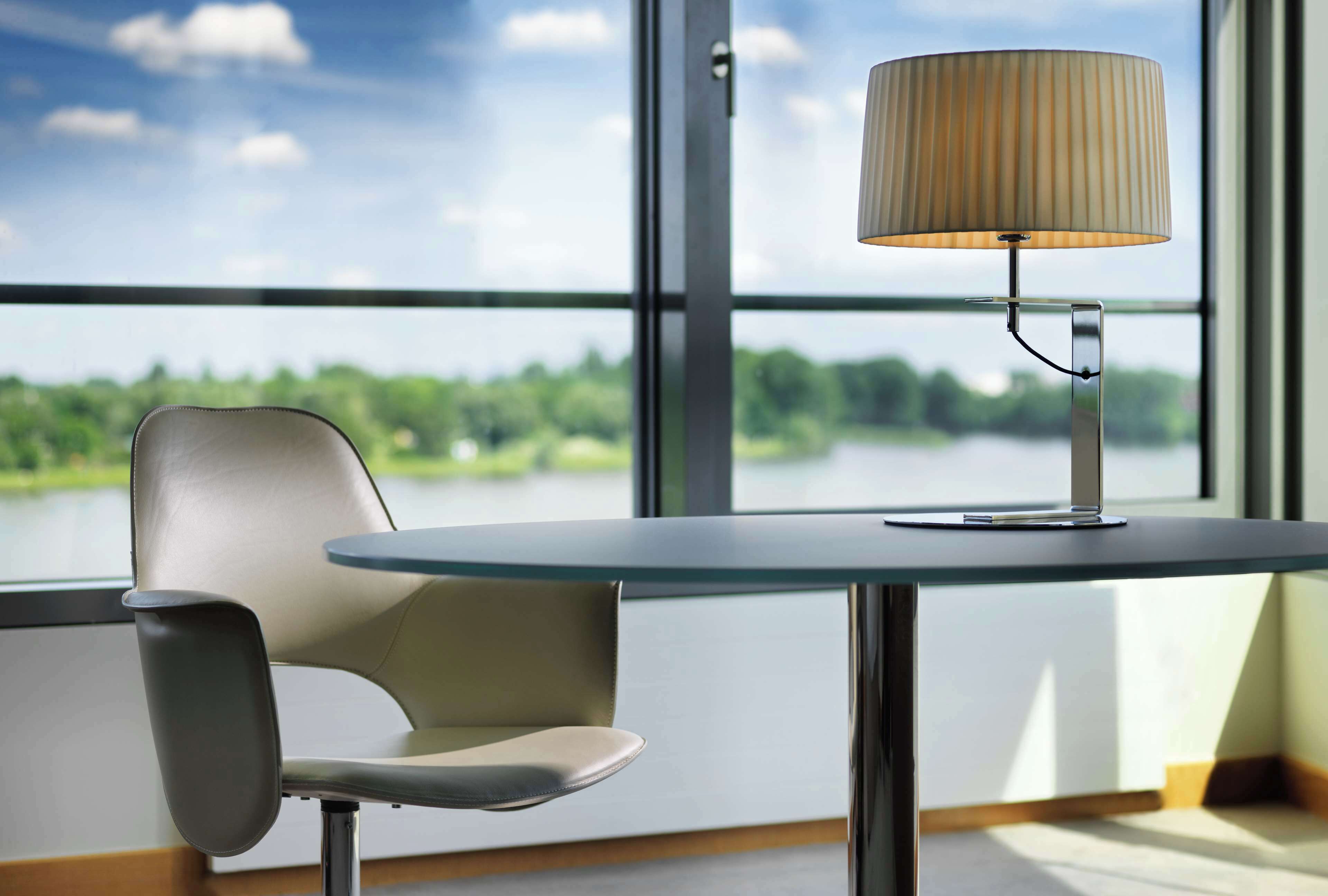 Una habitación moderna con una mesa, una silla, una lámpara y un ventanal con vistas a un lago y árboles.