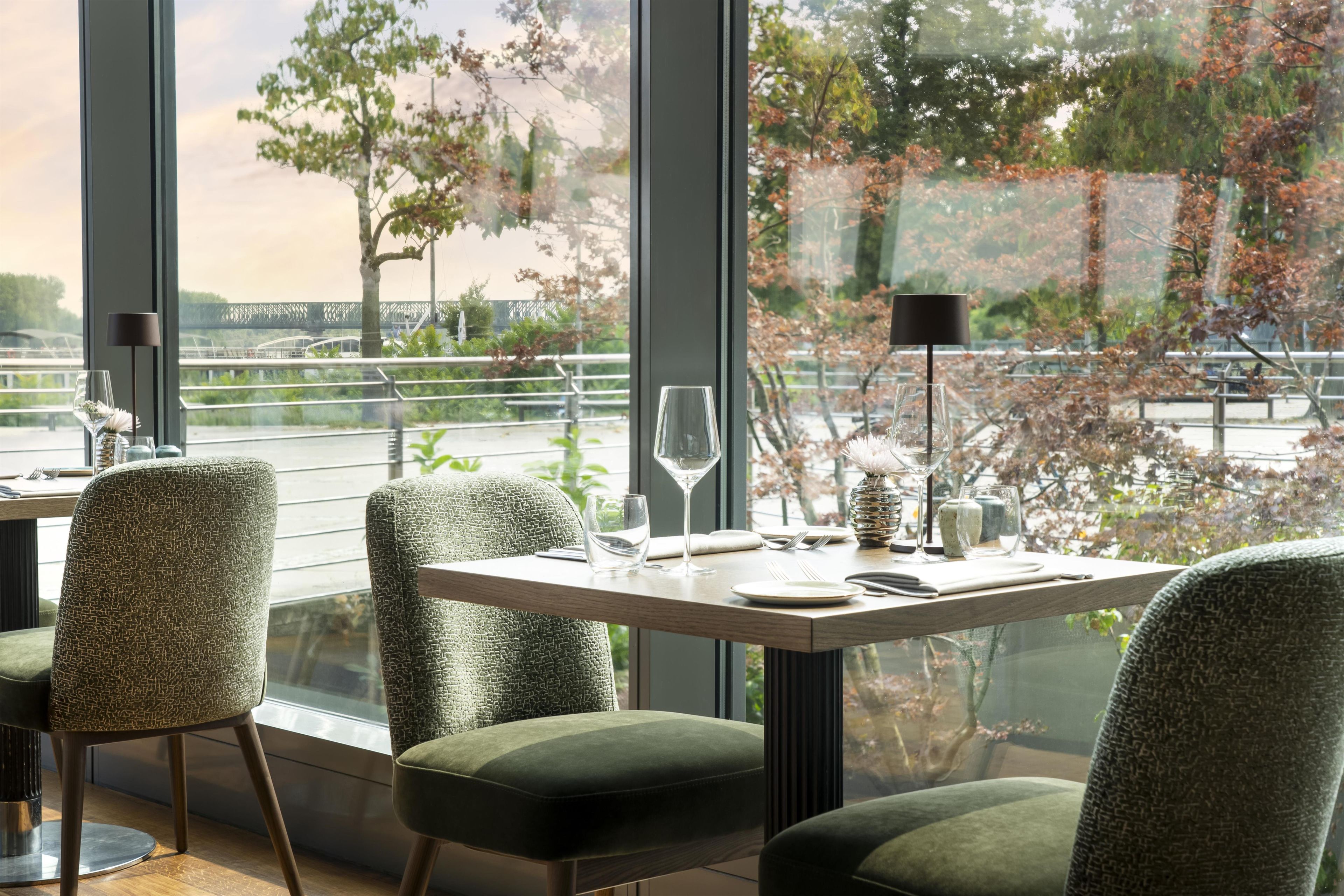 A dining area with tables set for two, featuring green upholstered chairs and a view of trees through large windows.