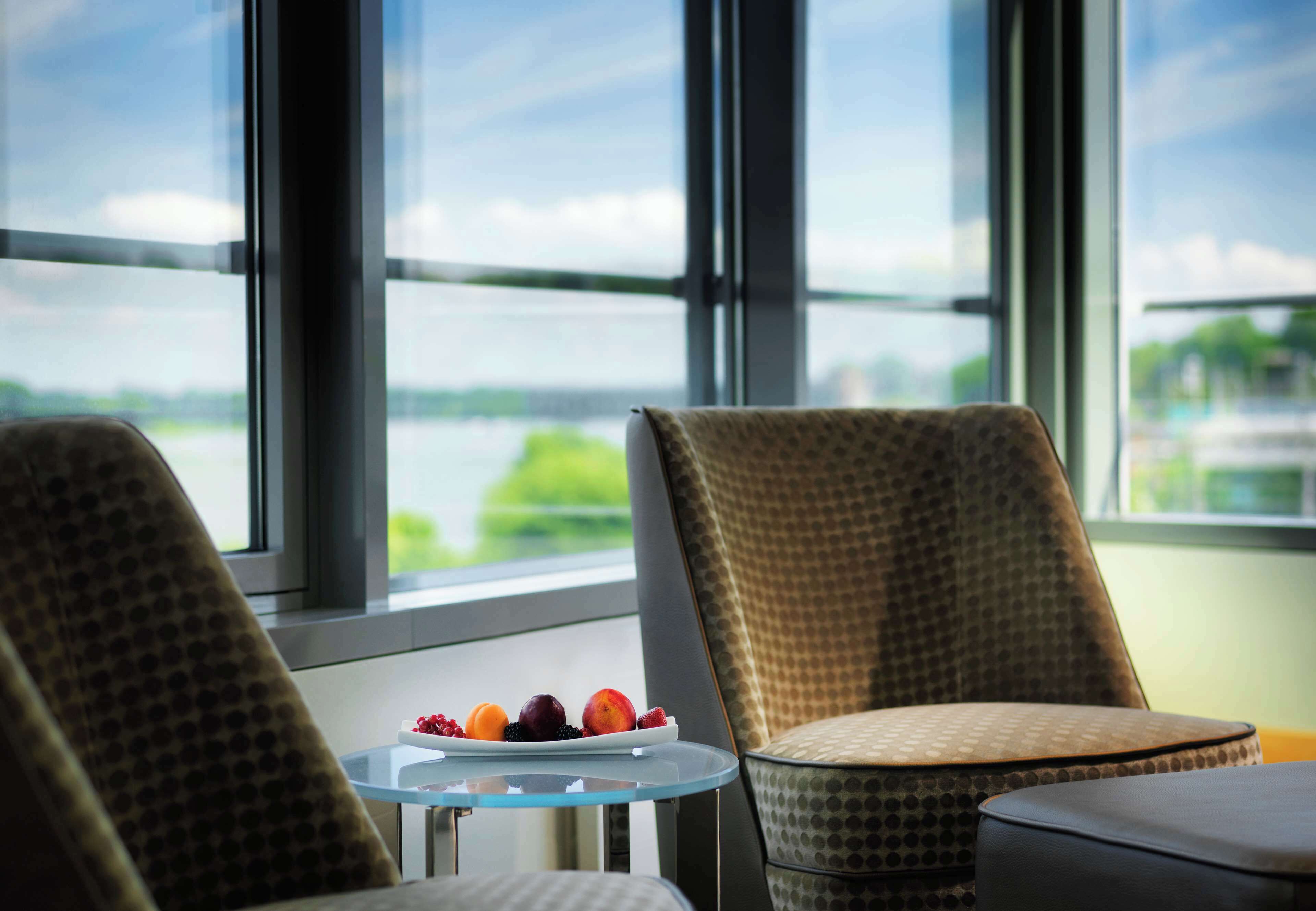 Ein Zimmer mit Blick auf ein Gewässer, große Fenster, ein kleiner Tisch mit Obstschale und zwei stilisierte Stühle.