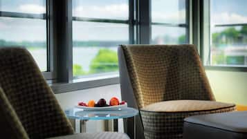 Ein Zimmer mit Blick auf ein Gewässer, große Fenster, ein kleiner Tisch mit Obstschale und zwei stilisierte Stühle.