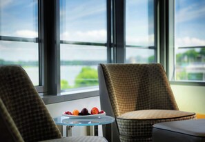 A room with a view of a body of water through large windows, a small table with a fruit bowl, and two patterned chairs.