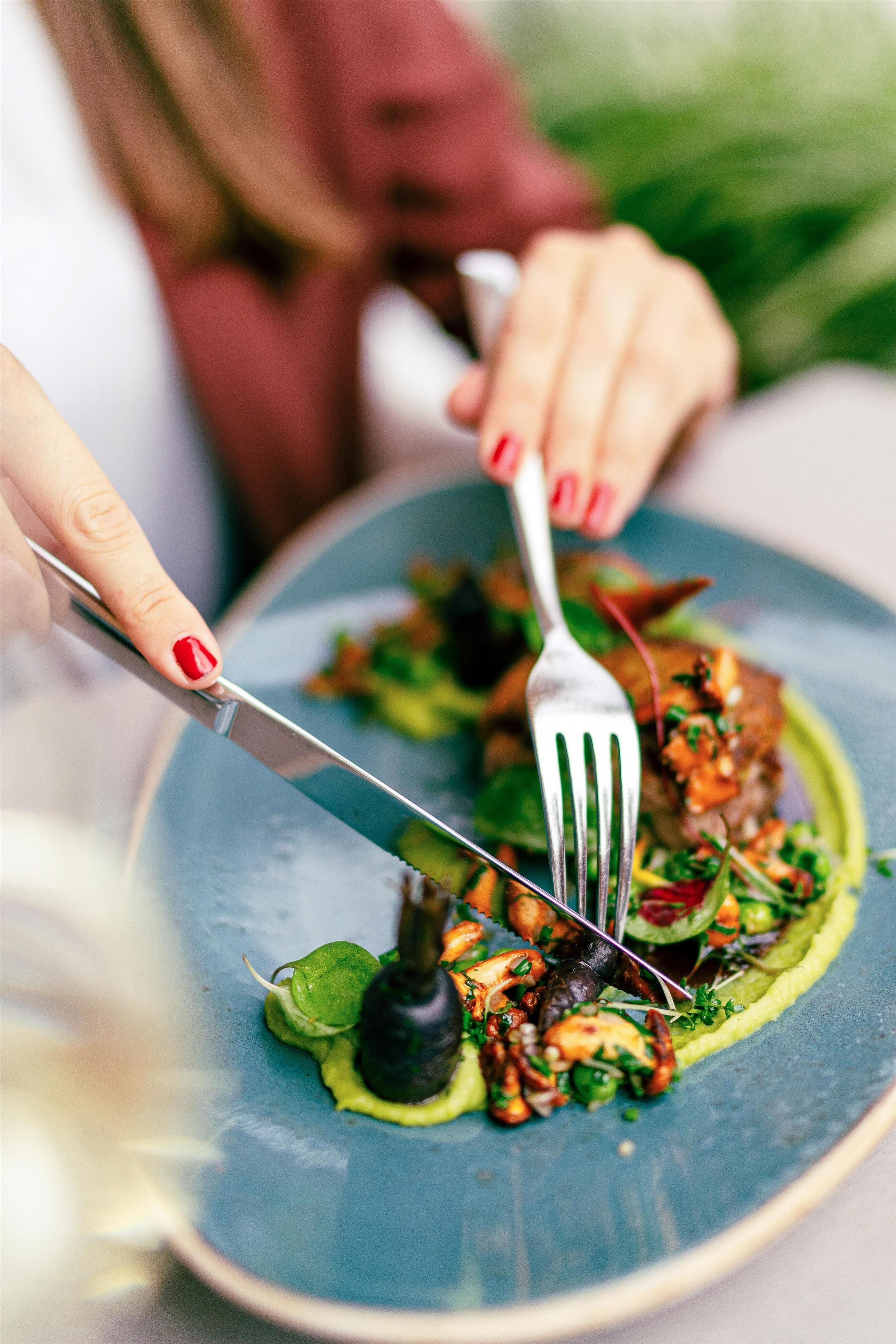 A person holding a fork and knife, about to eat a dish with greens and a dark sauce.