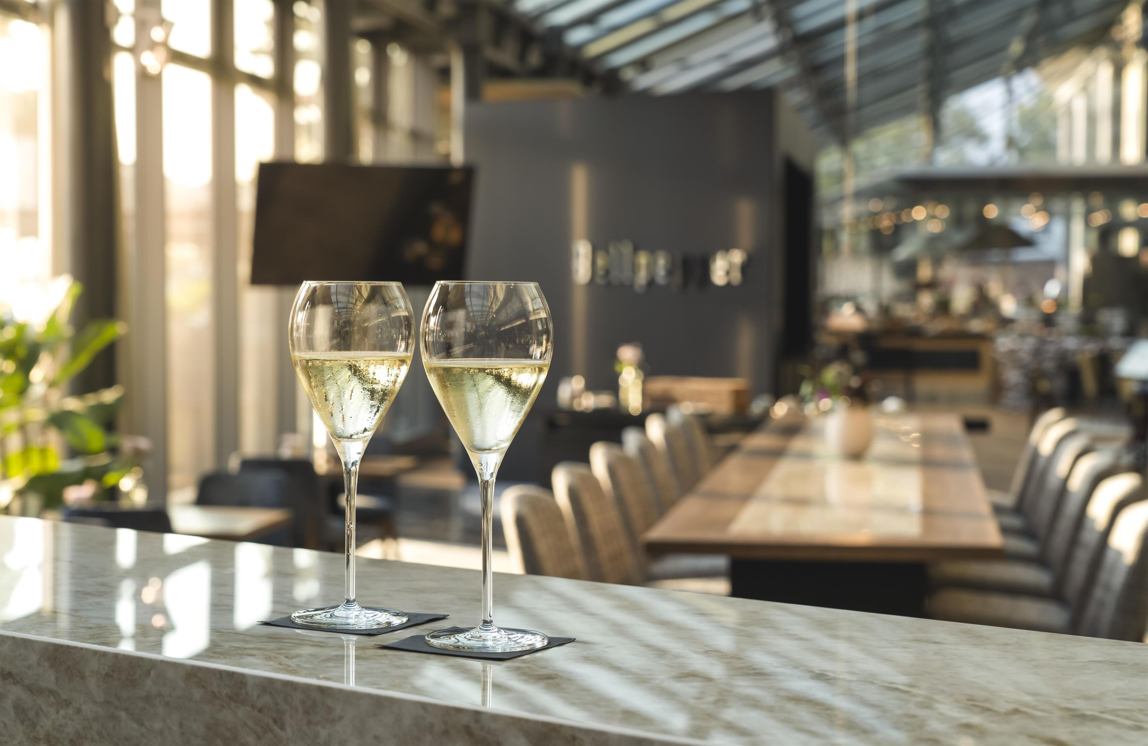 Two glasses of white wine on a marble bar counter in a modern restaurant with a long dining table and chairs.