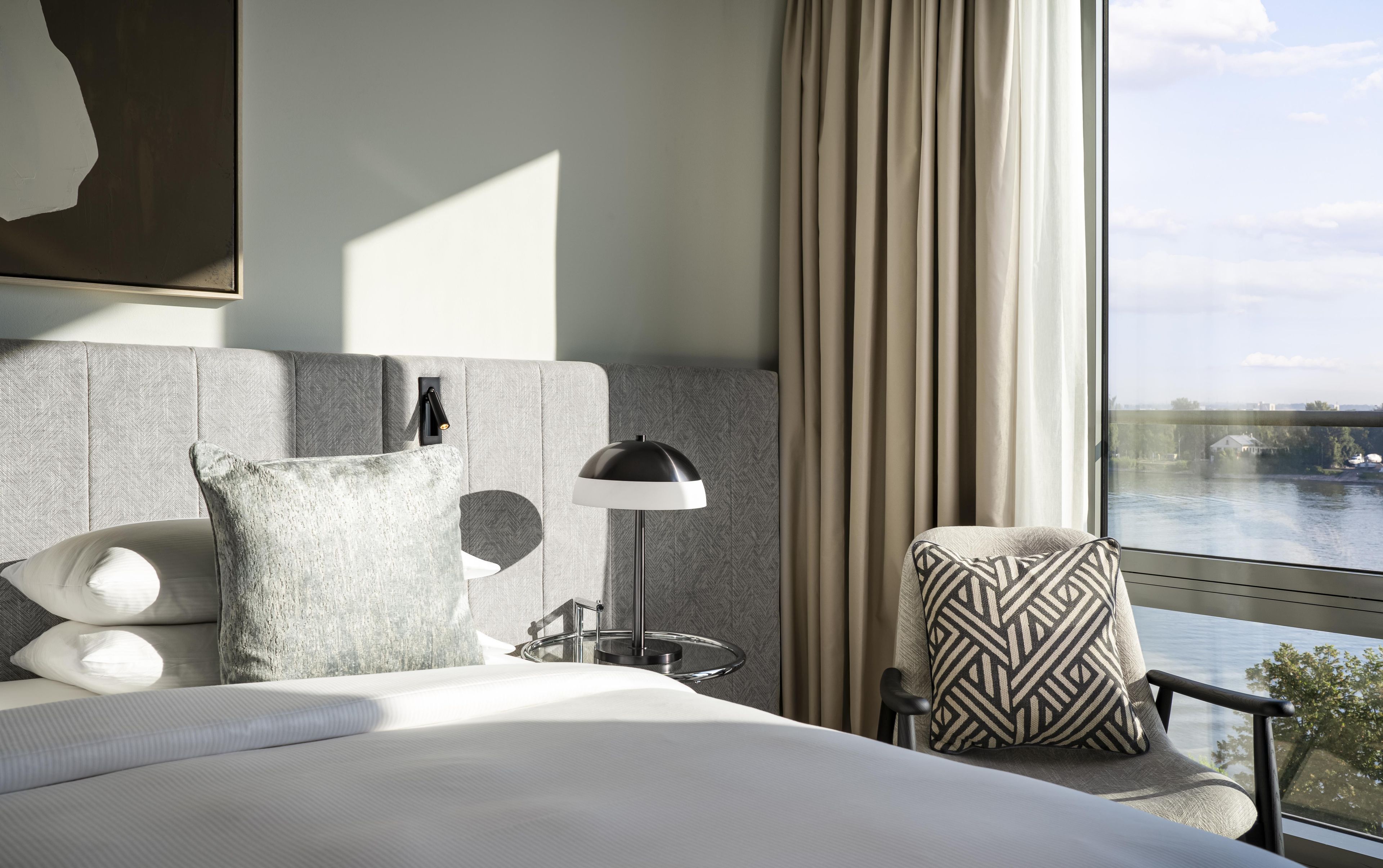 Habitación de hotel con una cama, una mesita de noche con lámpara, una silla y vista al río.
