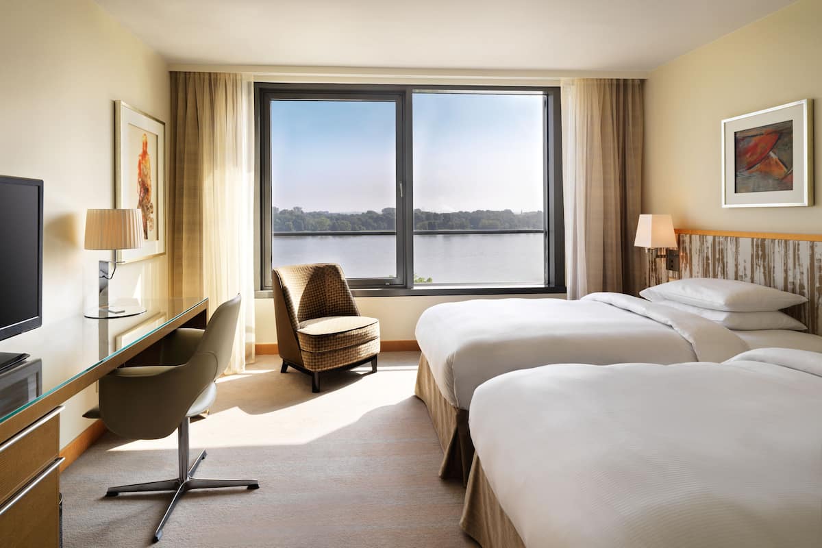 A hotel room with a large window, a desk with a TV, two beds, and a chair.
