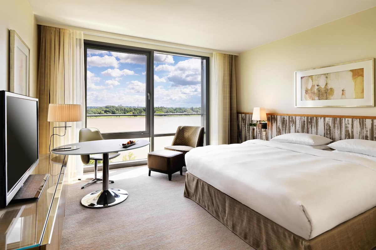 A hotel room with a large bed, a desk with a TV, a chair, and a view of a body of water.