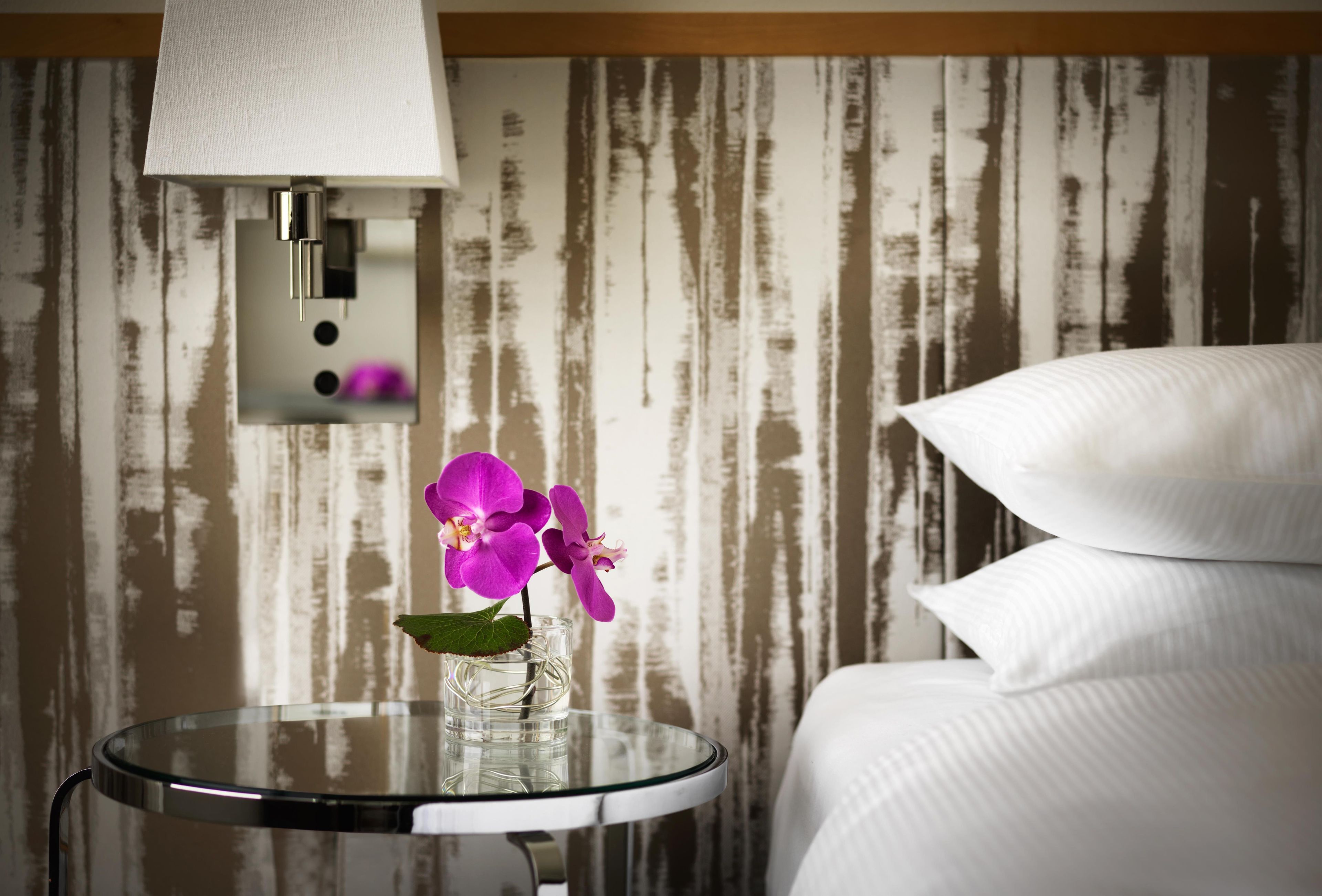 Une pièce comprenant un lit, une table ronde en verre ornée d’un vase de fleurs violettes et une lampe fixée au mur.