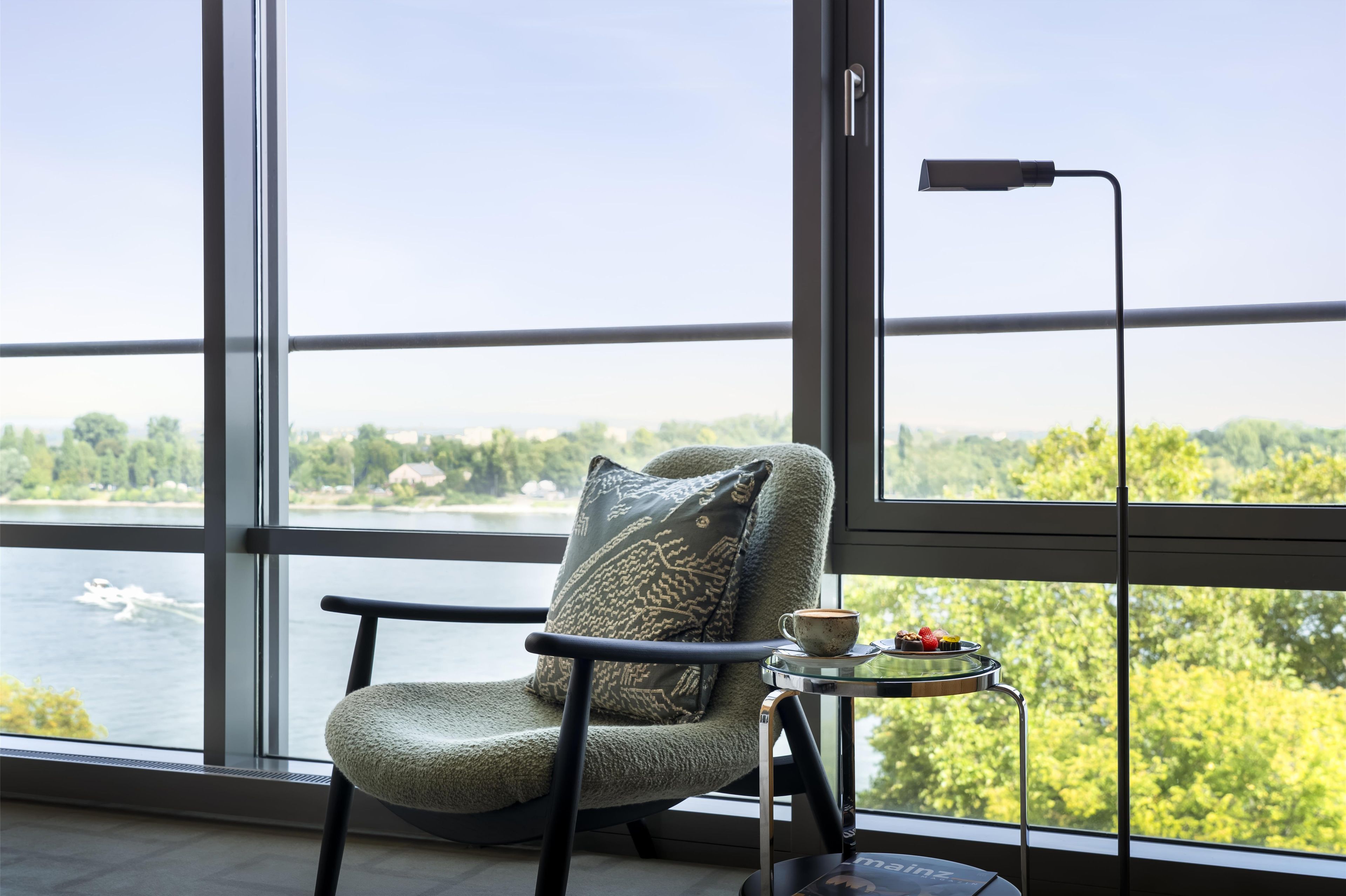 Ein Sessel mit Kissen und ein kleiner Tisch mit Tasse und Snacks, platziert neben einem großen Fenster mit Blick auf Wasser und Grünflächen.