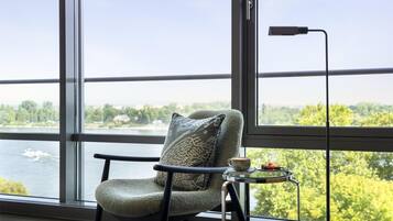 Ein Sessel mit Kissen und ein kleiner Tisch mit Tasse und Snacks, platziert neben einem großen Fenster mit Blick auf Wasser und Grünflächen.