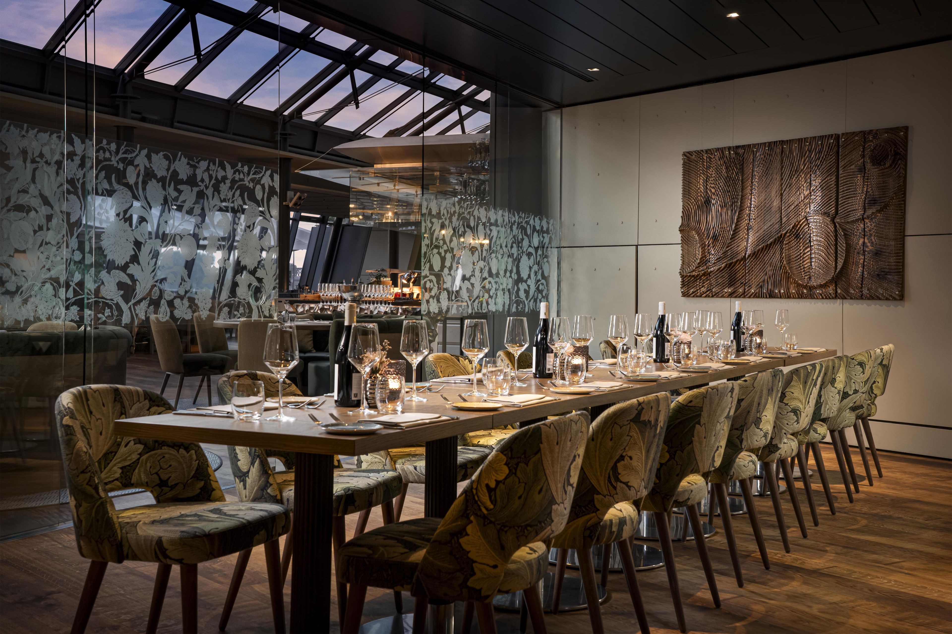 A dining area with a long table set for a meal, surrounded by patterned chairs, and a large abstract artwork on the wall.