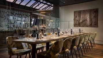 A dining area with a long table set for a meal, surrounded by patterned chairs, and a large abstract artwork on the wall.