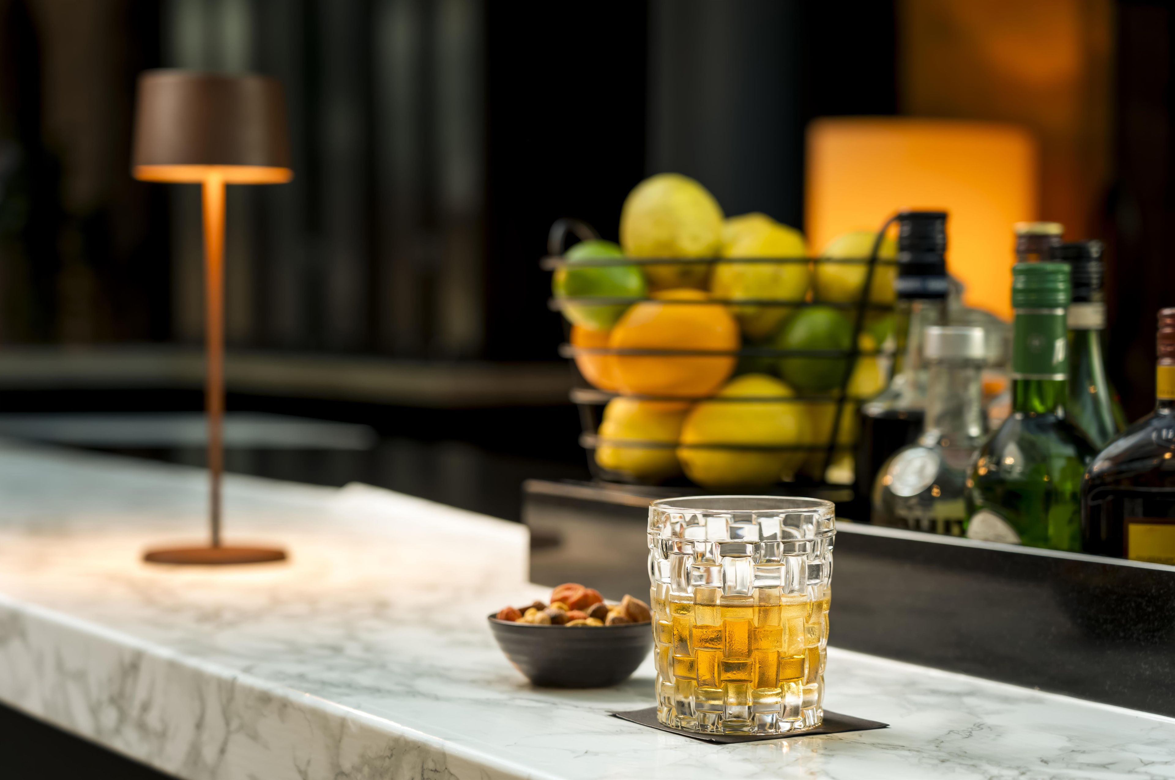 A bar area with a glass of amber liquid, a bowl of nuts, and a fruit basket.