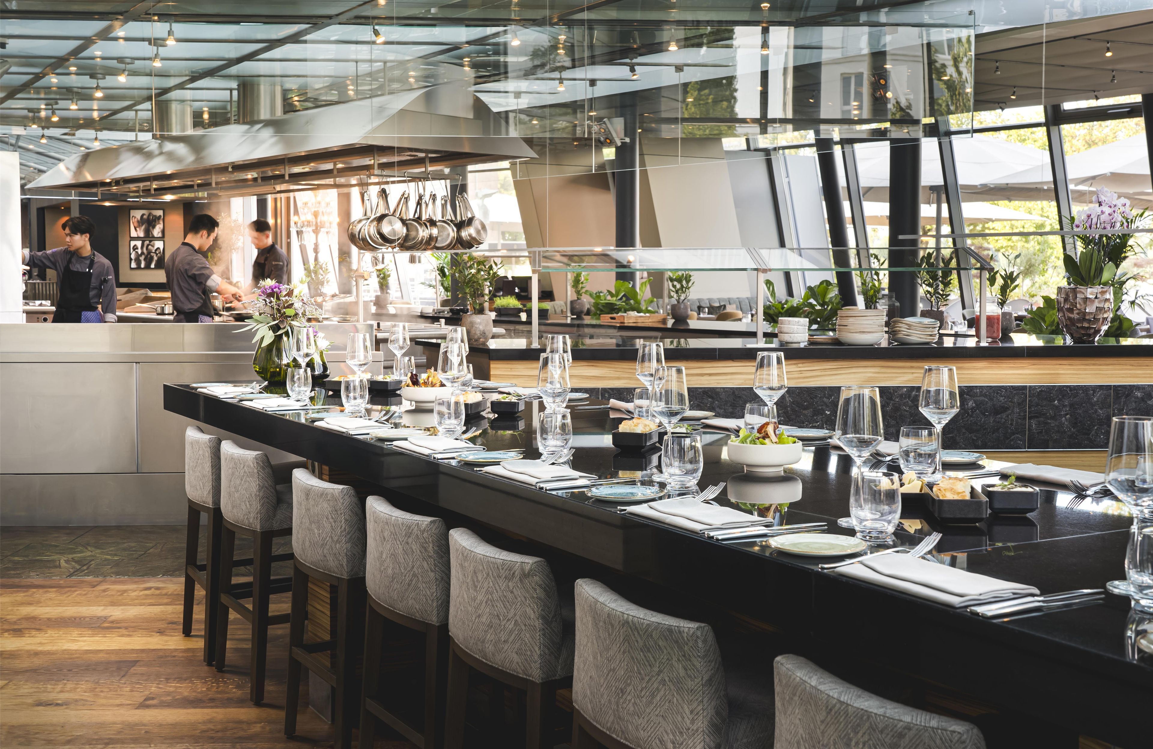 A modern restaurant with a long bar area, neatly set tables, and a kitchen in the background.